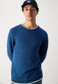 Blå strikkegenser med rund hals, med et teksturert mønster og kontrastfarget hvit kanting ved halsen og nederste del.