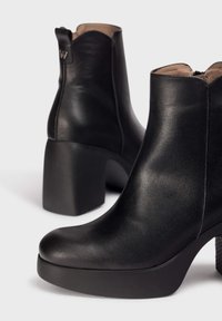 Botas de tobillo de cuero negro con un alto tacón de bloque, punta redondeada y una plataforma texturizada. Presenta un cuello festoneado y cremallera lateral.