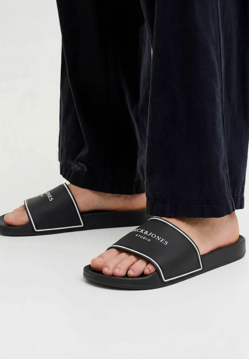 Pieds portant des sandales noires à enfiler avec un liseré blanc et le texte "JACK & JONES STUDIO", assortis à un pantalon noir.