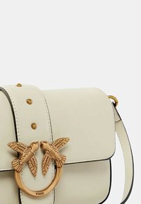 Bolso de cuero blanco con un cierre en forma de pájaro dorado, con un diseño plano y dos pájaros decorativos. Presenta ribetes negros y tachuelas doradas decorativas.