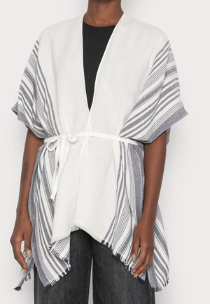 Persona vistiendo un kimono blanco y gris con rayas, con cinturón, sobre una blusa negra y pantalones oscuros, de pie frente a un fondo liso.