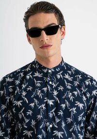 Camicia a maniche lunghe blu navy con motivo a foglie bianche, dotata di colletto a bottoni e bottoni frontali. Gli occhiali da sole neri completano il look.