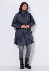 Giacca grigia impermeabile con un motivo astratto nero, dotata di cappuccio, zip frontale e tasche laterali; abbinata a leggings grigi e stivali neri.
