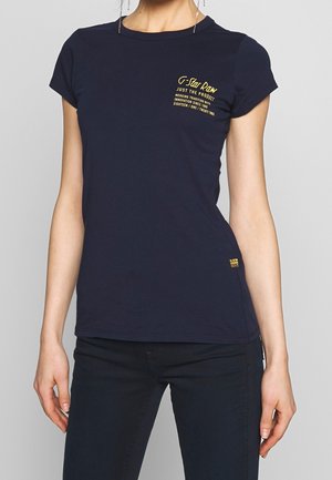 Mujer que lleva una camiseta entallada azul marino de G-Star Raw con texto de logo amarillo en el pecho y pantalones negros, frente a un fondo gris liso.