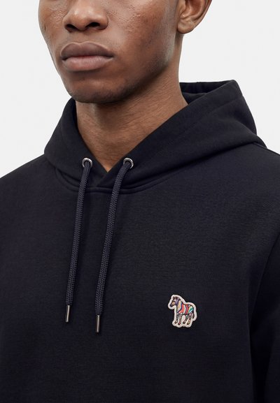 Gros plan d'une personne portant un sweat à capuche noir avec un petit logo zèbre rayé multicolore sur la poitrine et des cordons noirs avec embouts métalliques.