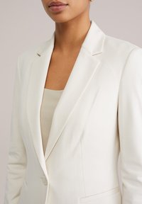 Moteris dėvi kreminės spalvos švarkelį ant šviesiai smėlio spalvos dryžuoto megztinio, matoma nuo kaklo iki vidurio liemens, fone – vienspalvis fonas.