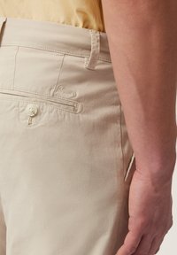 Bež pamuk hlače s ravnim stražnjim džepom, kopčanjem na dugme i izvezenim detaljem. Glatka tekstura i krojeni fit, prikazujući minimalistički dizajn.