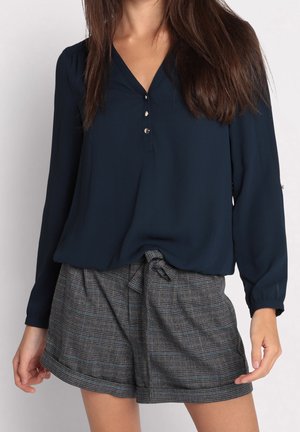 Vrouw draagt een marineblauwe blouse met lange mouwen en knopen, gecombineerd met hooggetailleerde grijze geruite korte broek, vastgebonden met een stoffen riem.