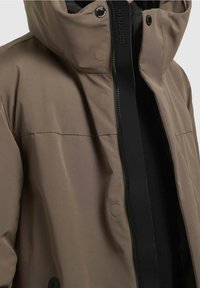Chaqueta impermeable marrón con cuello alto, cremallera frontal, botones a presión y detalles en negro. Presenta una textura suave y un diseño minimalista.