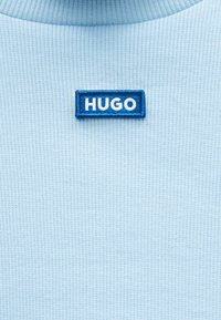 Ύφασμα σε ανοιχτό γαλάζιο με ριγέ υφή έχει ένα ορθογώνιο γαλάζιο κεντητό ένθετο με τη λέξη "HUGO" σε λευκά γράμματα.