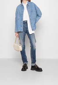 Chaqueta de mezclilla en azul claro con botones al frente, camisa blanca debajo, jeans azules y zapatos de plataforma negros con acentos dorados, sosteniendo un bolso crema.