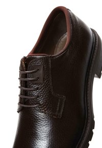 Zapato de cuero marrón con acabado texturizado, punta redondeada y diseño de cordones. Presenta un forro marrón en contraste y mínimos acentos de metal.