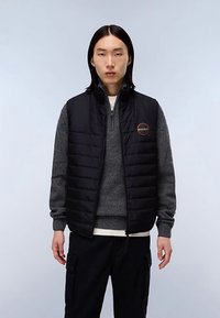 Černá prošívaná vesta s vysokým límcem, nošená přes šedý svetr. Má kulaté logo na hrudi, hladkou látku a zapínání na zip.