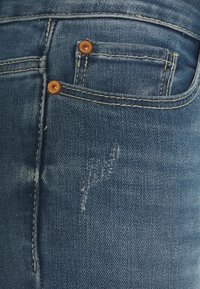 Vaqueros de mezclilla azul con un ligero desgaste, costuras doradas, dos remaches de cobre en el bolsillo frontal y detalles de abrasión visibles.