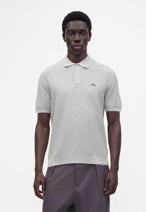 Jeune homme portant un polo gris clair à manches courtes avec un petit logo vert sur la poitrine gauche et un pantalon foncé.