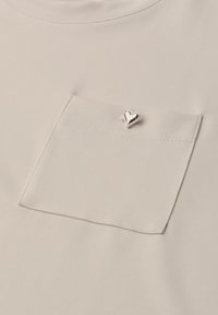 Poche en tissu beige avec une petite épingle brillante en forme de cœur argenté fixée près du centre supérieur de la poche.