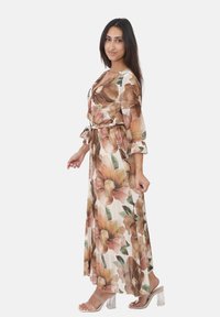 Lang blomstret kjole i beige, med store brune og pink blomster, lange ærmer, volumiøse manchetter og et bånd i taljen. Gennemsigtige hælede sandaler.