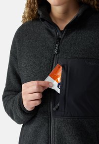 Dark gray fleece jacket with a black panel and zippered chest pocket. A hand reaches into the pocket, retrieving a packet with orange accents.