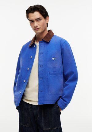 Mand iført en blå skjortejakke med brun krave, beige skjorte under og mørke denimbukser, poserende med hænderne i lommerne.