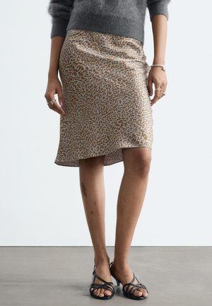 Femme portant une jupe beige et marron imprimé léopard longueur genou, un pull gris, des sandales à talons noires à brides, ainsi que des bagues et un bracelet en argent.