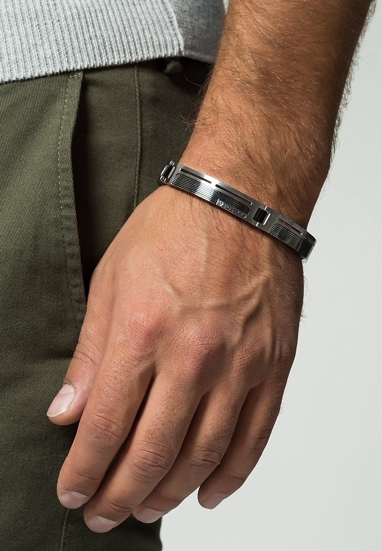 Main masculine à la peau claire portant un bracelet en métal argenté, reposant près d'un pantalon vert olive et d'une manche de pull en maille gris.