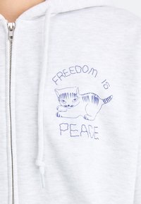 Hallatav hall zip-up kapuutsiga sviiter, millel on sinise tikandiga tekst "VABADUS ON" kassi illustratsiooni kohal ja "RAHU" all. Pehme kangast tekstuur, lõdvestunud disain.