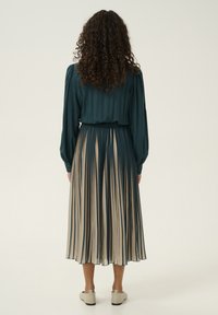 Dark green blouse with long sleeves, elastic waist cream and dark green pleated skirt, ending mid-calf, paired with light-colored slip-on shoes.