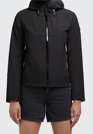 Vrouw draagt een zwart capuchonjack met rits en donkere denimshorts, staand met ontspannen armen langs haar zij.