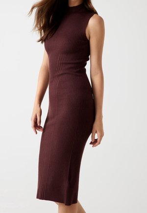 Femme portant une robe midi en maille côtelée sans manches de couleur brun foncé, debout devant un fond uni gris clair.