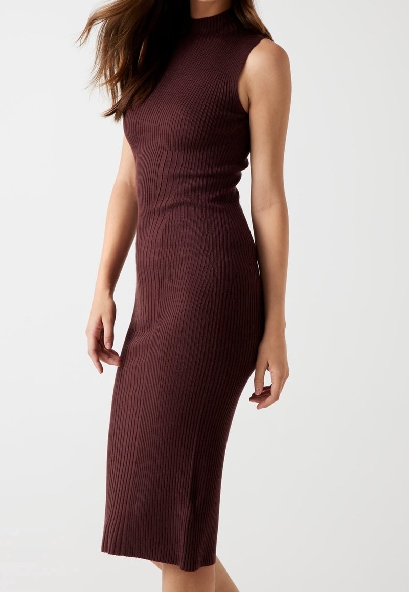 Femme portant une robe midi en maille côtelée sans manches de couleur brun foncé, debout devant un fond uni gris clair.