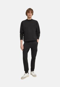 Sudadera negra con puños acanalados, combinada con pantalones de chándal negros que tienen un bolsillo lateral, y zapatillas blancas con detalles en gris.