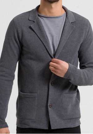 Mężczyzna w teksturowanej, ciemnoszarej marynarce zapinanej na guziki, na jasnoszarej koszulce, trzymający prawą ręką klapę marynarki.