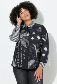 Sort skjorte med hvide, fremtrædende mønstre, herunder sol- og måne-designs. Stribede ærmer og krave. Blødt stof, knappes foran.