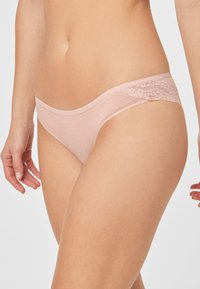 Lichtroze strings met een zachte stof en kanten accenten aan de zijkanten, gepresenteerd tegen een witte achtergrond.