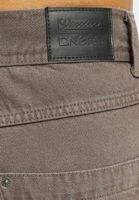Jeanșii din denim maro au o suprafață texturată, cusături detaliate și o etichetă din piele neagră cu inscriptionarea "Denim by DNGRS".