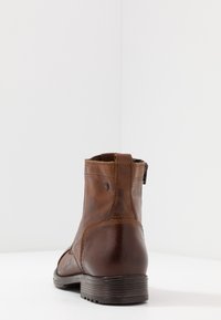 Bottine en cuir marron avec fermeture éclair sur le côté, coutures visibles et semelle marron foncé texturée, vue de dos sur une surface blanche.
