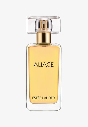 Klar glasflaske med rektangulær form, gyldne detaljer og firkantet låg. Indeholder en gul væske mærket "ALIAGE Estee Lauder."