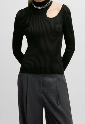 Mujer con un top negro de manga larga con canalé y un corte asimétrico cerca del hombro, y pantalones sueltos gris oscuro.