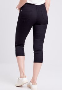 Schwarze Capri-Leggings aus dehnbarem Stoff, mit Gesäßtaschen und glatter Oberfläche, kombiniert mit weißen Sneakers.