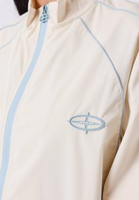 Lichte crème jasje met lichtblauwe accenten, een hoge kraag en een ritssluiting aan de voorkant. Bevat een blauw logo op de linkerborst.