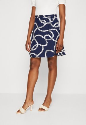 Kvinde iført en mørkeblå nederdel med hvidt rebmønster, taljen bundet, kombineret med beige flettede højhælede sandaler og hvid top.