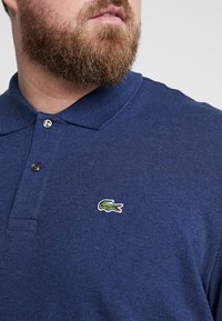 Marineblaue Polohemd aus strukturiertem Stoff, mit einem Knopfleisteneinsatz mit zwei Knöpfen und einem grünen Krokodil-Logopatch auf der linken Brust.