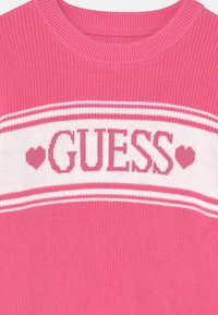 Suéter de punto rosa con una franja horizontal blanca que lleva la palabra "GUESS" en rosa, acentuada por pequeños corazones a cada lado.