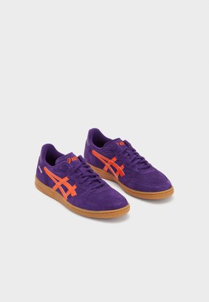 Paire de baskets en daim violet avec des rayures et logos orange, semelles en caoutchouc marron, et fermeture à lacets sur fond blanc.