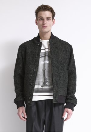 Jonge man draagt een zwarte, gestructureerde jas met knopen over een wit T-shirt met print en zwarte broek, staand tegen een witte achtergrond.