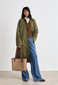 Olive green long coat with gold buttons over a brown cable-knit sweater, paired with wide-leg blue jeans and a brown patterned tote bag.