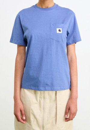 Person iført en kortærmet blå t-shirt med brystlomme og beige nederdel med lynlås foran, stående mod en hvid baggrund.