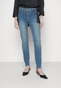 Světlé modré džíny s úzkým střihem, s elastickým pasem, předními kapsami a kontrastním prošitím. Spárováno s černými sandály na podpětku.