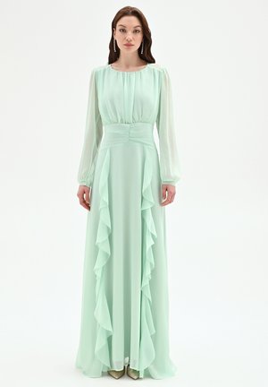 Donna in piedi che indossa un lungo abito verde menta con maniche lunghe di pizzo e balze a cascata sul davanti, abbinato a orecchini pendenti.