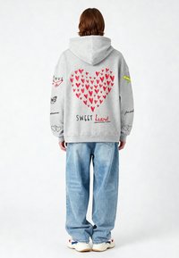 Sudadera gris con gráfico de corazón rojo y acentos coloridos, combinada con jeans anchos de color azul claro y zapatillas blancas. Elementos de texto presentes.